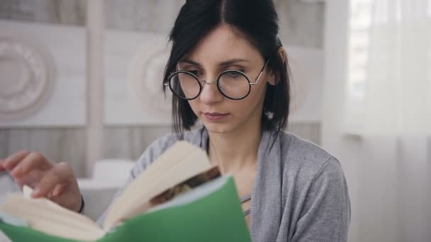 Uma jovem de óculos senta-se na cama em casa e folheia um livro. Uma menina bonita lê um livro interessante — Vídeo de Stock