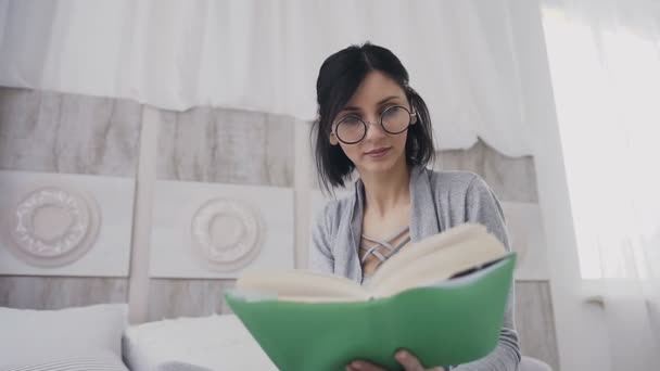 Beyaz kız yataktan kitap okumak rahatlatıcı bir gözlük. Yatağında bir kitap okuma gözlüklü genç güzel esmer kadın. Sabah, duygular kavramı — Stok video