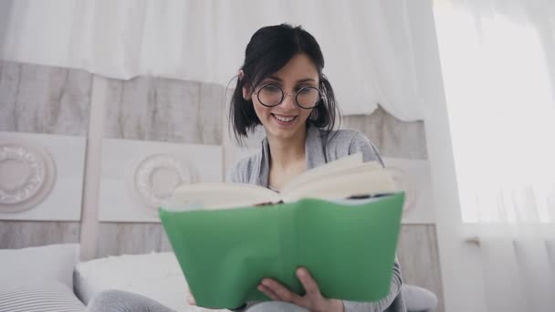 Средний план. Красивая ученица в очках читает забавную книгу дома. Девушка улыбается, читая книгу на кровати в белой спальне. Утренний свет, концепция эмоций — стоковое видео