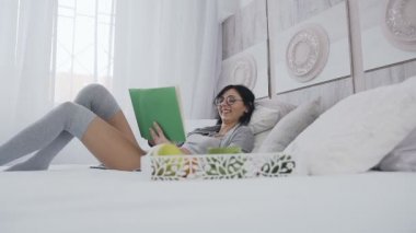 Güzel esmer yatak odalı evde yatakta yatarken, o ilginç bir kitap okurken gülümsüyor. Sabah, lezzetli breakfast, duygu ve öğrenme kavramı