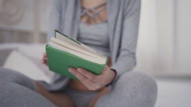 Yakın çekim. Güzel esmer kız bardaklarda yatakta oturur ve bir kitap okuma için tutar. Genç bir kadın ilginç bir kitap evde yeşil bir kapak açılır.