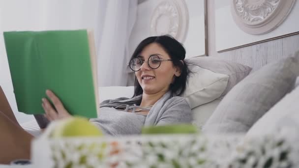 Primer plano. Hermosa morena en gafas acostada en la cama en el dormitorio de su casa, sonríe mientras lee un libro interesante. Mañana, delicioso desayuno, concepto de emociones y aprendizaje — Vídeo de stock