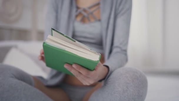 特写。美丽的黑发女孩在眼镜坐在床上, 并持有一本书阅读。一个年轻女子在家里的绿色封面上打开一本有趣的书。 — 图库视频影像