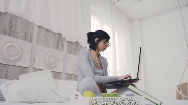 Mulher morena atraente jovem sentada na posição de lótus na cama no quarto branco com laptop escuro. Menina bonita em óculos digitando texto em um laptop em casa no quarto — Vídeo de Stock