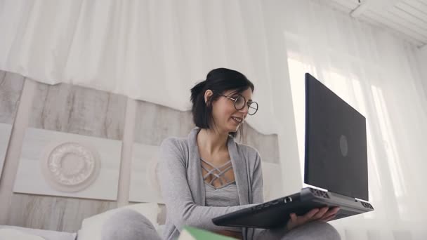 Eine hübsche Frau mit Brille tippt Text auf einem Laptop und sitzt auf einem Bett in einem gemütlichen weißen Schlafzimmer. eine junge Frau mit dunklen kurzen Haaren kommuniziert mit Freunden im Internet in sozialen Netzwerken auf einem Gadget — Stockvideo