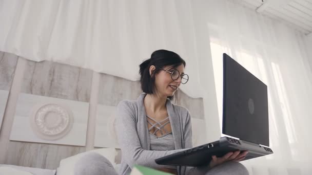 Een mooie vrouw in glazen typen van tekst op een laptop die zittend op een bed in een gezellige witte slaapkamer. Een jonge vrouw met kort donker haar communiceert met vrienden in sociale netwerken op een gadget op het Internet — Stockvideo