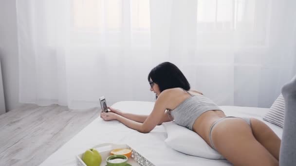 Uma garota bonita e atraente em óculos está deitada na cama em uma lingerie cinza e meias longas e faz foto de selfie no telefone. Isto está a acontecer numa sala branca brilhante. Ela está sorrindo e — Vídeo de Stock
