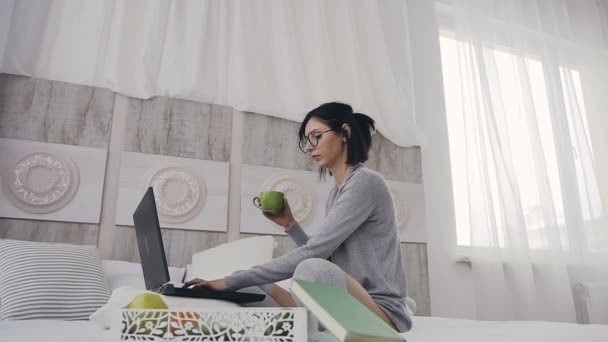 Fröhliches Mädchen mit Brille, das am Laptop tippt, um soziale Medien zu teilen, im Bett sitzt und morgens zu Hause Kaffee trinkt — Stockvideo