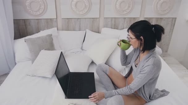 Vacker ung kvinna med laptop pc, dricka varmt te med citron medan du sitter på sängen i de vita sovrummet — Stockvideo