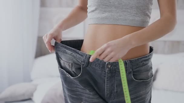 Close-up. A slim girl is measuring her waist with a meter tape. She is also checking how much her jeans are loose after she has lost a bit of weight — Stock Video