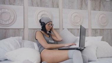 Pretty genç kız Amerikalı kadın siyah kulaklık ve müzik, dans ve beyaz oda yatakta otururken onu laptop çalışma gri şapka. Bir kız bardaklarda