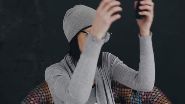 Una encantadora joven morena con sombrero gris y gafas de sol escucha música en los auriculares y canta, sobre un fondo negro — Vídeos de Stock