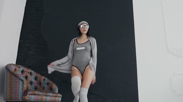 Mladá brunetka v sluneční brýle poslouchá hudbu v její černé sluchátka a tancem poblíž barevné křeslo na pozadí černé a bílé zdi, vnitřní — Stock video
