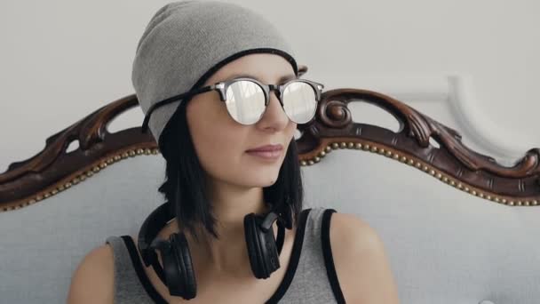 Portrait of beautiful young girl wearing headphones and sunglasses looking sideways and smiling in indoor. On the background of a white wall — Stock Video