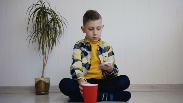 Knap kereltje, zittend op de vloer in de buurt van de witte muur thuis met behulp van een mobiele telefoon en drinkwater uit een rode kop — Stockvideo