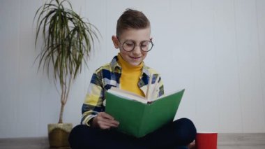 Güzel küçük okullu çocuk komik beyaz kapalı yerde otururken bir kitap okuma gözlük. Evde komik kitap okumak. Çocuk ilginç ders kitabı okuyun