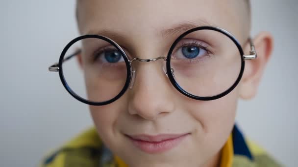 Gros plan sur le visage d'un beau garçon en lunettes avec de beaux yeux bleus qui regardent dans la caméra. Le petit garçon a les yeux bleus et porte des lunettes élégantes. En fond blanc — Video