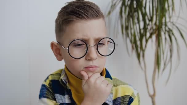 Close-up. Retrato do pensamento infantil masculino. O estudante com olhos azuis vestindo óculos é concebido olhando na direção e meditando em algo. Em fundo branco, interior — Vídeo de Stock