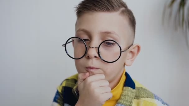 Expressive little 7-8year child with a beautiful smile and blue eyes with glasses is thinking about an interesting idea, looks in the side ways with a smart face and holds his beard with his hand — Stock Video