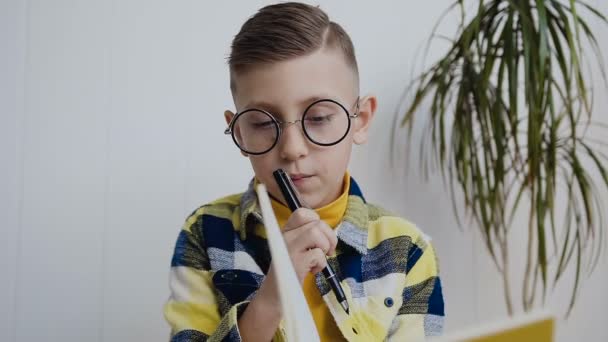 Un chico de escuela elegante con gafas se sienta en el fondo de una pared blanca y hace notas interesantes en un cuaderno. Un niño concebido hace notas en un libro de texto amarillo. En interiores — Vídeos de Stock