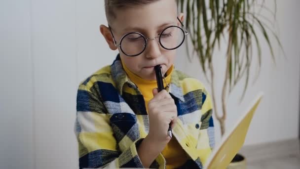 Egy elegáns iskola fiú szemüveg ül a háttérben, egy fehér fal, és teszi érdekes jegyzetek a notebook. Fogant fiú sárga tankönyv teszi megjegyzések. Beltéri — Stock videók