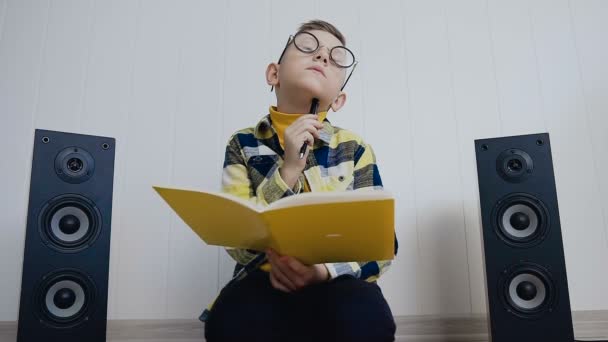 Blond chłopiec w okularach z podręcznika siedzi na podłodze w domu i rejestruje coś tam ciekawe w notebooku. Uczeń ma pracę, że został poproszony w szkole robi w domu — Wideo stockowe