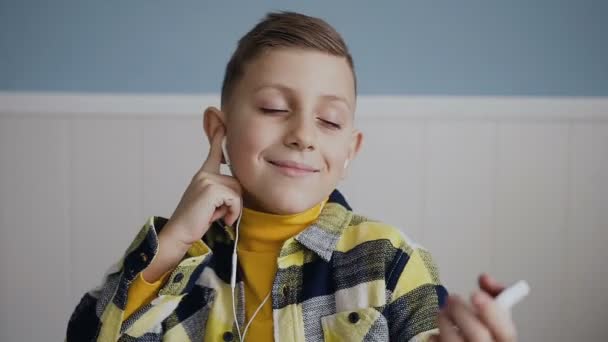 Retrato. Un hermoso chico rubio de 7-8 años escuchando música en los auriculares blancos, bailando asintiendo con la cabeza. De cerca. En fondo blanco, en interiores — Vídeos de Stock