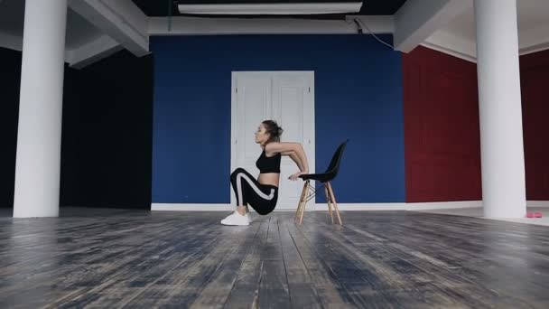 Sports girl performs exercises with squatting leaning hands around on the chair. A young fitness woman trains performing exercises for her legs. Healthy way of life, workout. She dressed in black — Stock Video