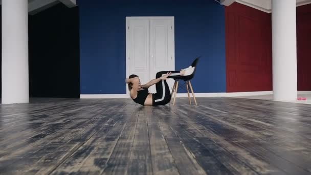 Chica de fitness haciendo ejercicio de prensa acostado en el suelo apoyado contra la silla en el gimnasio. Ella se vistió con ropa deportiva negra — Vídeos de Stock
