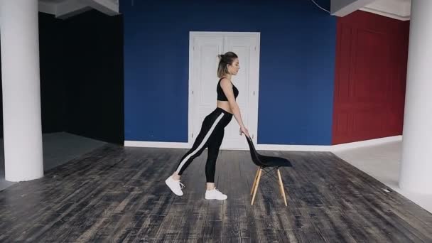 Fitness jeune femme vêtue de vêtements de sport sombres s'entraîne au gymnase seul. Elle fait l'un des types de pompes en s'accrochant à la chaise. Se maintenir fort et en bonne santé — Video