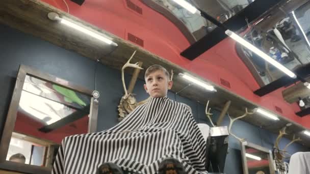 Вид снизу. Ребенок делает прическу в мужском парикмахерском салоне. Парикмахер замачивает расческу в воде для расчесывания волос. Мальчик сидит в кресле в парикмахерской и парикмахер делает ему новый — стоковое видео