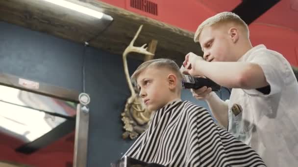 Fryzjer z dzieckiem klienta w salonie fryzjerskim. Za pomocą trymera i grzebień fryzjerski w fryzjera. Słodkie dziecko siedzi na krześle w salon fryzjerski. Nowa fryzura — Wideo stockowe
