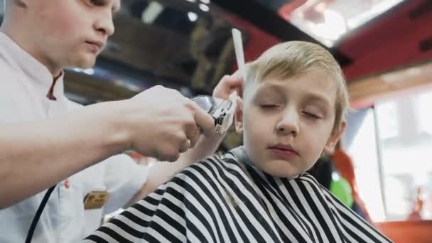 Kuaför berber içinde küçük bir çocuk için şık bir saç kesimi yapıyor. Sihirli küçük çocuk mens güzellik salonunda yeni bir saç modeli yapmak — Stok video