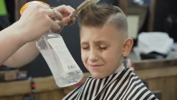 Der Friseur spritzt Wasser aus dem Zerstäuber auf den Kopf der kleinen Kundin im Friseursalon, die auf das Ende des Haarschnitts wartet — Stockvideo