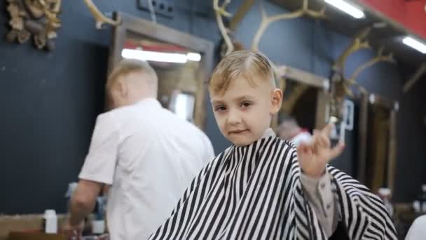 Un niño pequeño se sienta en una silla en una barbería mirar a la cámara y muestra con la mano que todo está bien y le gusta su barbero. Bonito peinado. Niño en el salón de peluquería para hombre — Vídeos de Stock