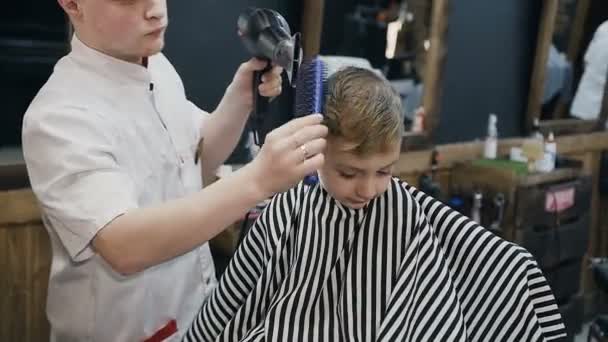 Cabeleireiro profissional secagem cabelo de menino com secador de cabelo e escova na barbearia. O menino de cabelo molhado seca-se pelo secador de cabelo. Barbearia — Vídeo de Stock