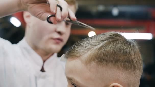 Парикмахер делает укладку волос на коротких волосах. Милый мальчик стрижется у парикмахера, сидя в кресле в парикмахерской — стоковое видео