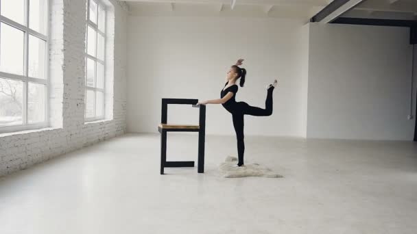 Rzemieślnik gimnastycy, nagrzewa się w szkole sport. Ładna dziewczyna praktyk gimnastyka w białym domu, ona ciągnie się, podziały, stojąc na jednej nodze, a i opierając ręce na krześle. Trening w — Wideo stockowe