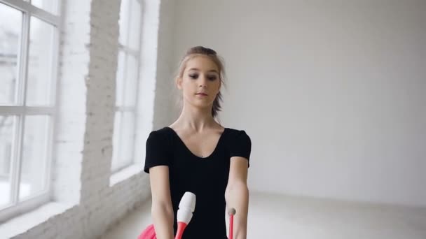 Rítmica bela ginasta segurando macas faz movimentos acrobáticos no fundo branco no salão de ginástica. Menina vestida de corpo preto e saia rosa — Vídeo de Stock