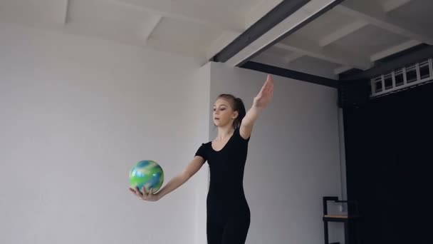 Hermosa chica rubia vestida con traje de cuerpo deportivo negro realiza bailando con pelota en la escuela de gimnasia. Gimnasta con la pelota en sus manos haciendo movimientos acrobáticos en el deporte indoor en el blanco — Vídeo de stock