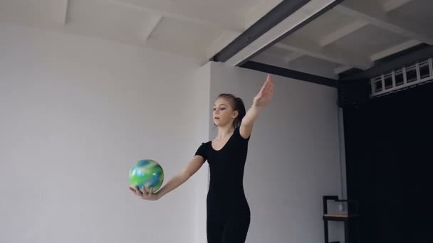 Gyönyörű szőke lány öltözött fekete sport test alkalmaz hajtja végre, táncos torna iskola labdát. A labdát a kezével csinál akrobatikus tornász mozdulatok-on a fehér beltéri sport