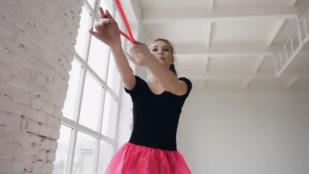 Όμορφη γυμνάστρια εκμετάλλευση εθιμοτυπικά μπαστούνια κάνει ακροβατικές κινήσεις σε άσπρο φόντο στην Γυμναστικού αίθουσα. Κοπέλα ντυμένη στα ροζ φούστα και μαύρο σώμα — Αρχείο Βίντεο