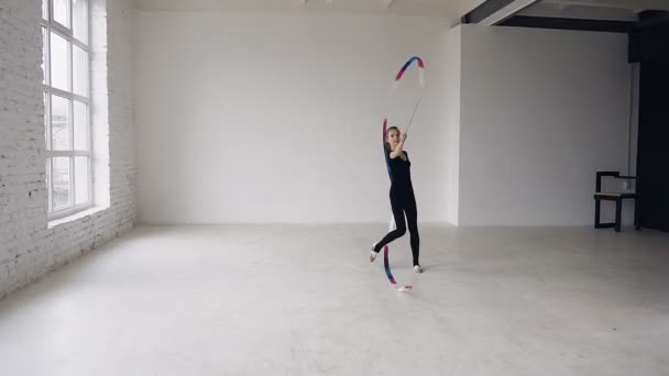 La gimnasta rítmica sostiene la cinta en una mano y la agita y baila. Ella se vistió de cuerpo negro y se está preparando para la actuación en una competición deportiva, mirando a una cámara — Vídeo de stock