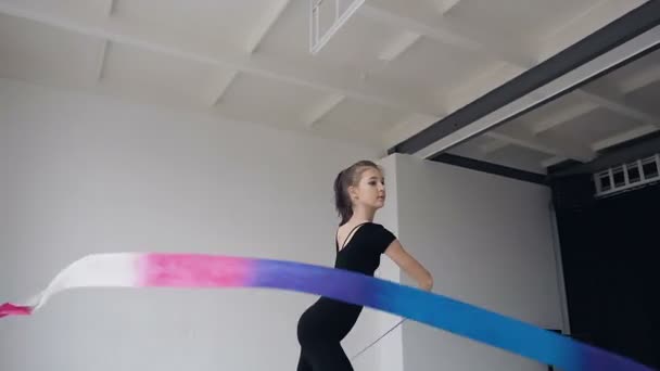 Jolie gymnaste féminine dansant avec ruban de couleur à l'école de gymnastique. La gymnastique fine dans le corps noir effectue l'exercice de gymnastique avec un ruban dans le studio blanc et pose à la caméra — Video