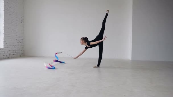 Little gymnastics in black body doing the gymnastics exercise with a colored ribbon in the white studio. Cute girl makes stretching exercise in sport gym — Stock Video