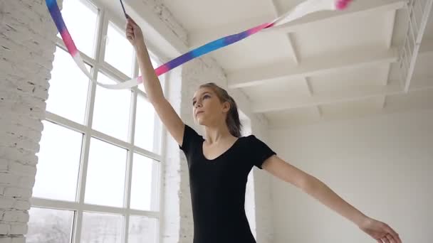 Ginasta bonito vestido com corpo preto com fita colorida em sua mão cria movimento bonito e gracioso em interior em fundo branco perto das janelas. Escola de ginástica ou ballet — Vídeo de Stock