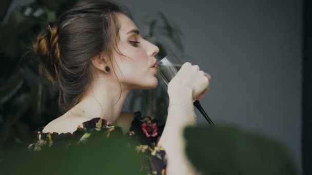Πλαϊνή όψη. Όμορφη νεαρή γυναίκα που πίνει σαμπάνια στο μπάνιο σε φόντο πράσινο λουλούδια. Όμορφη κοπέλα απολαμβάνει από την κατανάλωση της σαμπάνιας σε ένα καυτό λουτρό — Αρχείο Βίντεο