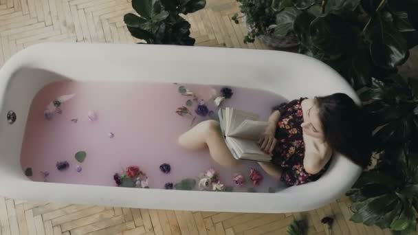 Una hermosa chica lee un libro y se relaja en un baño caliente lleno de leche y flores aromáticas. Mujer joven leer libro interesante en el baño — Vídeo de stock