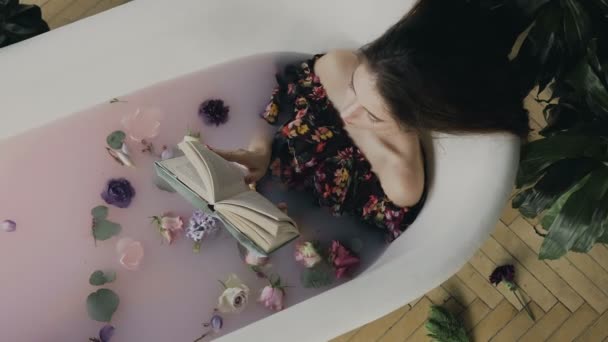 顶部视图。一个美丽的年轻女子读了一本书, 在温暖的浴缸里放满了牛奶和芬芳的花蕾。美丽的女人放松在牛奶浴躺在浴缸, 而阅读 — 图库视频影像