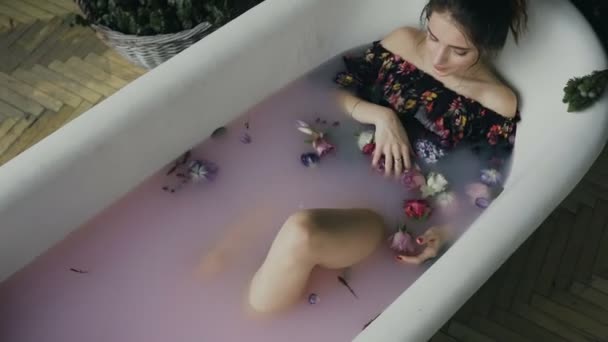 Söt ung kvinna avkopplande i mjölk bad med en rosa nyans och med många vackra doftande blommor efter en hård arbetsdag. Spa-behandlingar och hudvård — Stockvideo